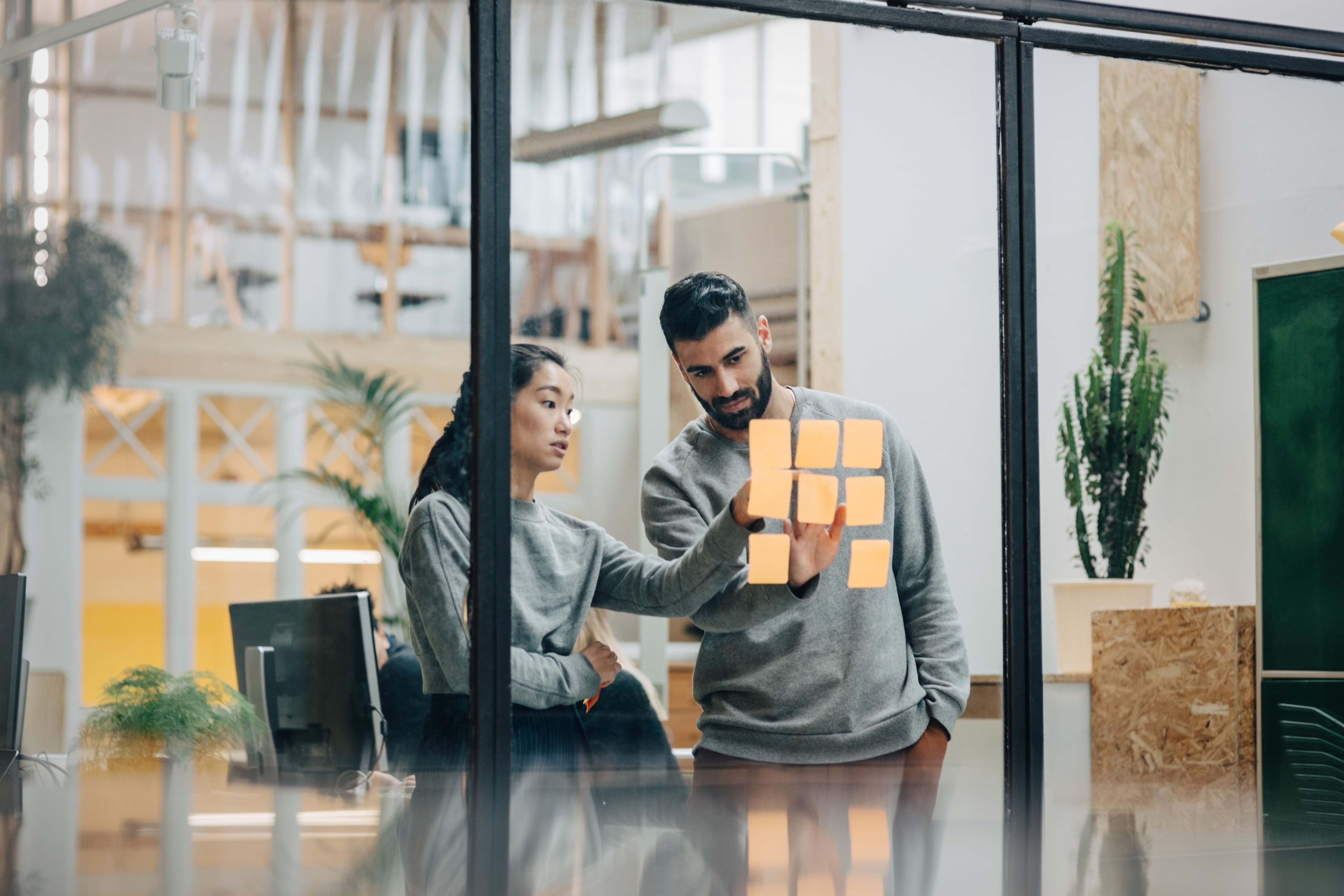 STOCK Businessman discussing with businesswoman sticking adhesive notes on glass wall in office.jpg